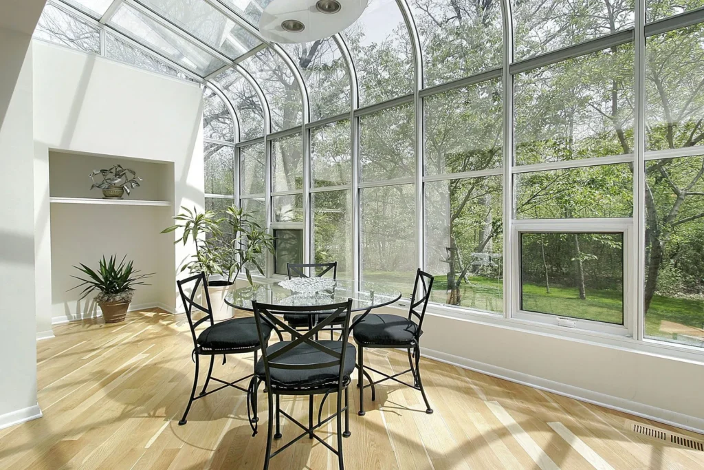 Conservatory style sunroom with curved glass roof and walls, light wood floor, and custom dining table installed by All Star Sunrooms.